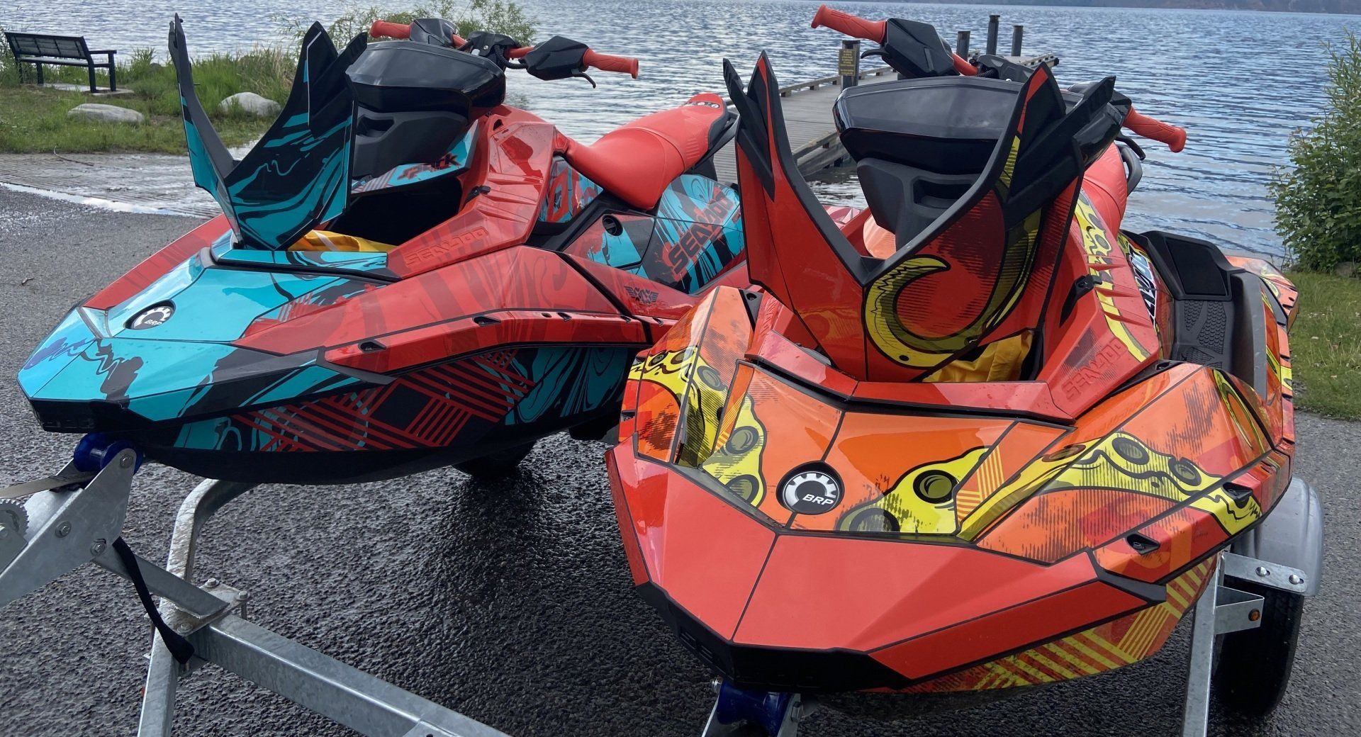 Two Sea-Doo Spark Trixx with red, orange and teal wraps on a trailer at a boat launch, the lake and dock behind them